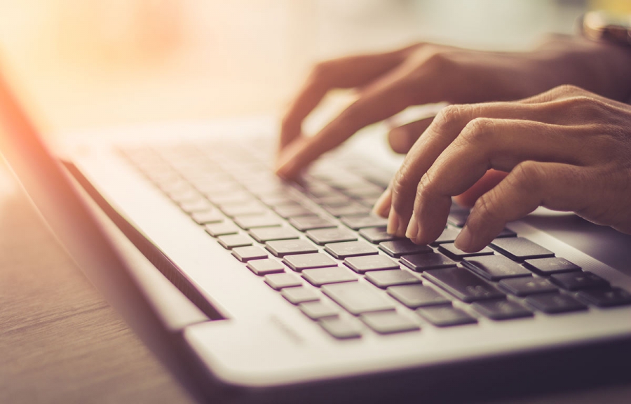 A person typing on a laptop keyboard with a blurred background and sunlight streaming in from the right side.