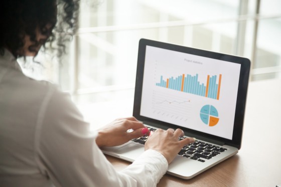 Woman in a white blouse working on a laptop with graphs on screen, in a modern office.