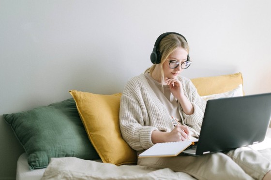 Woman sitting on bed with headphones, laptop, and notebook working on the side.