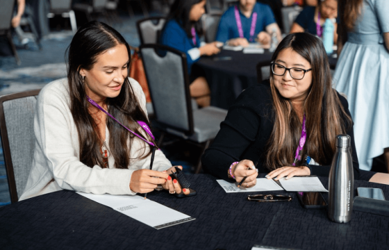 Two women sit at a table during a meeting, discussing and taking notes with a pen and paper.