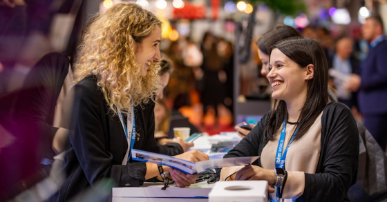 Two women chatting at a meeting with a blurred background of people and an event space.