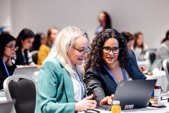 Two women working on a laptop in an event with people in the background and drinks on the table.