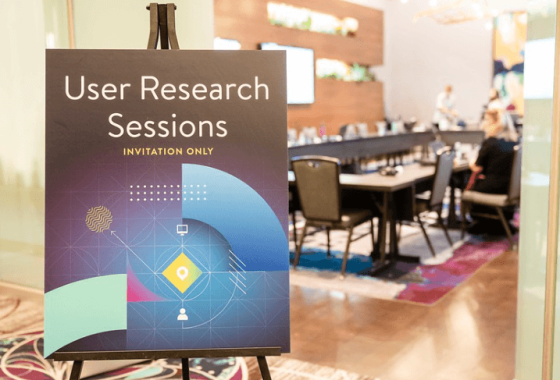 A User Research Sessions sign stands in front of a conference room with people sitting at tables and chairs.