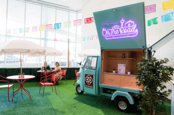 A food truck named "On the Road" sits in a building with people sitting at tables and umbrellas.