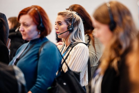 Woman with headphones and a microphone waits with a group of people in a blurred background.