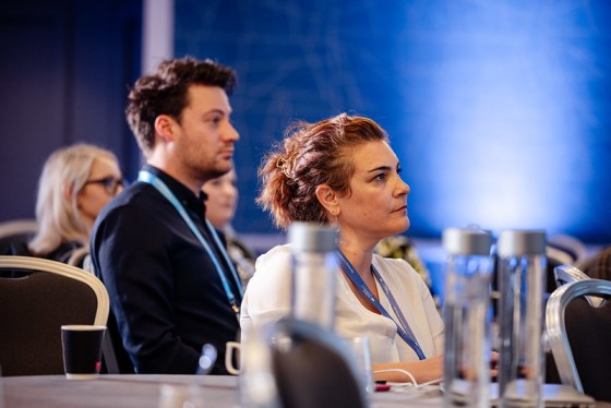 Audience listening intently in a conference room with water bottles and a blue backdrop in the background.