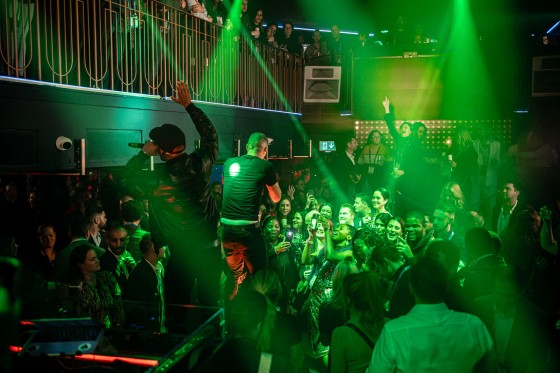 A crowd at an event with green lighting, and two people on stage with a microphone.