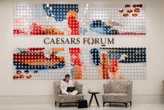 A woman is sitting in a chair in front of a large wall with Caesars Forum text on it.