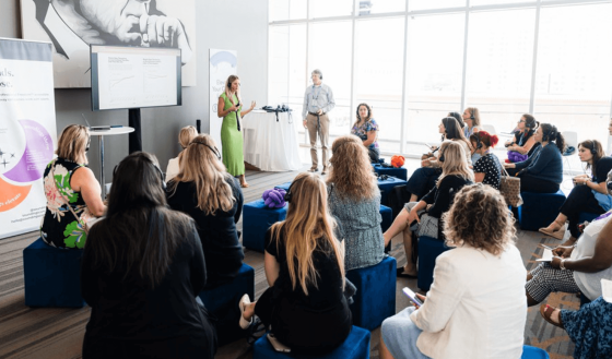 Seminar room with people in blue chairs listening to a woman in a green dress speak.