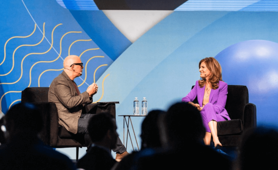 A man and woman are sitting on chairs while talking and there are people in front of them.