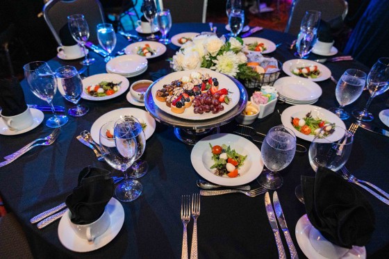 Table with black tablecloth and plates of food with napkins and water glasses set for an event.