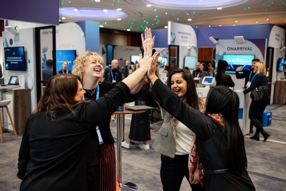 Four women high-fiving each other at a conference with a sign that reads "Onarrival" in the background.
