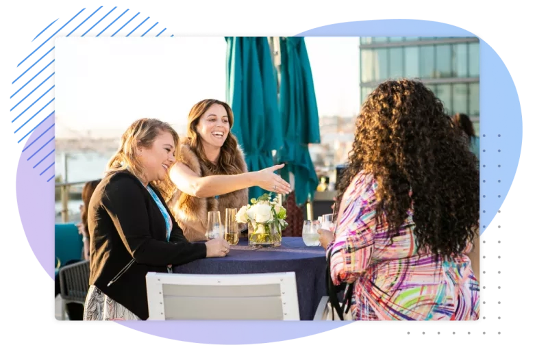 Three women at a table with drinks and flowers on it, laughing and talking in an outdoor setting.