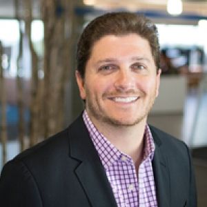 Brian Ludwig wearing a black coat and checkered shirt is smiling in front of a glass wall.