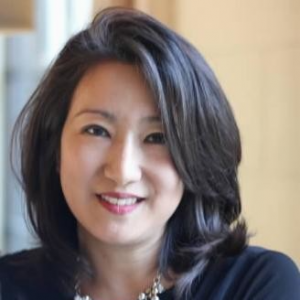 Portrait of Grace Lee, smiling and wearing a necklace, with a beige background.