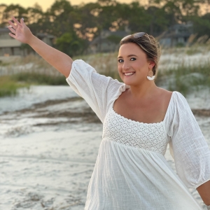 A woman in a white dress standing on a beach with her arm raised and smiling.
