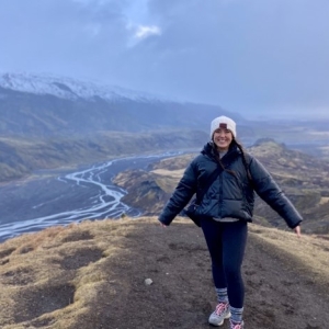 A woman stands on a mountain in Iceland wearing a hat and a jacket, with a river below her.
