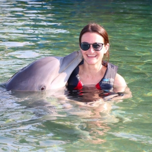 Woman in a life jacket in the water with a dolphin nuzzling her neck.
