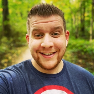 Greg Strong, a smiling man with a beard, wearing a t-shirt in a forest area.