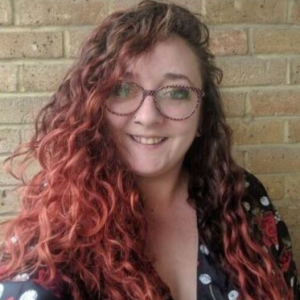 Woman with red hair wearing a black and white dress with glasses in front of a brick wall.