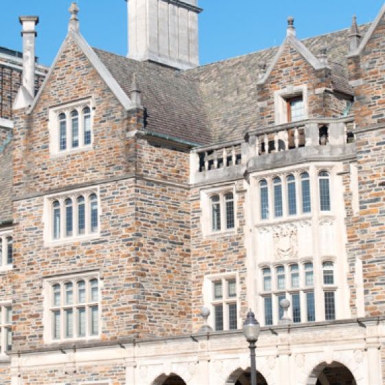 A brick building with many windows, a gray roof, and green trees in front of Duke University.