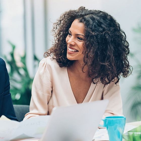 Two smiling people with documents and laptops in an office with plants in the background.