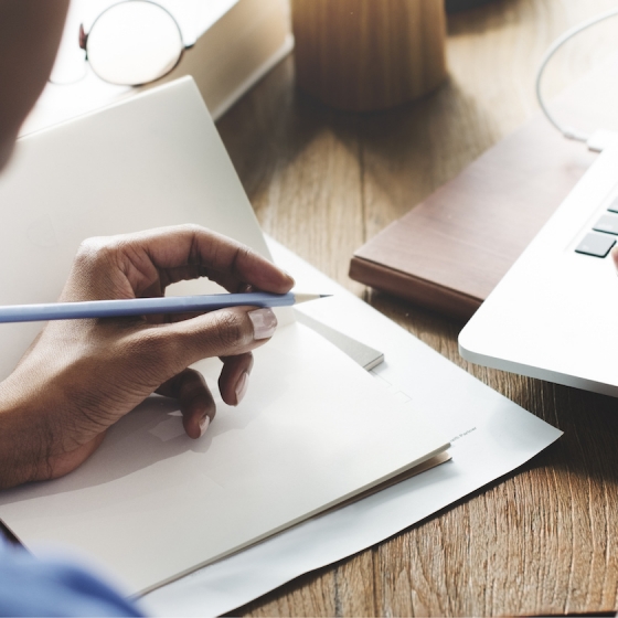 Person writing on paper and typing on laptop in a workspace with copy space to the right.