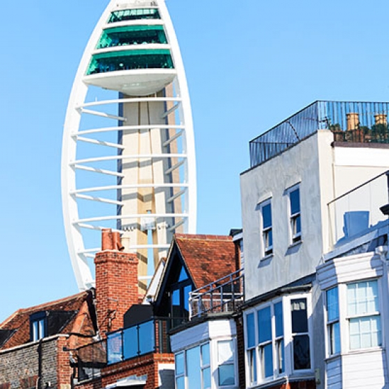 A tall tower, Spinnaker Tower, stands in front of buildings, with a clear blue sky above and a streetlight nearby.