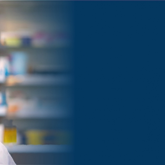 Pharmacy technician wearing a face mask and white lab coat with maroon shirt underneath in a pharmacy.