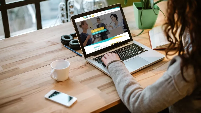 A woman using a laptop with the website open on a wooden table with a plant and coffee cup.