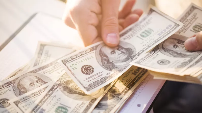 Hands holding a stack of $100 bills with the first bill in focus, showing George Washington.