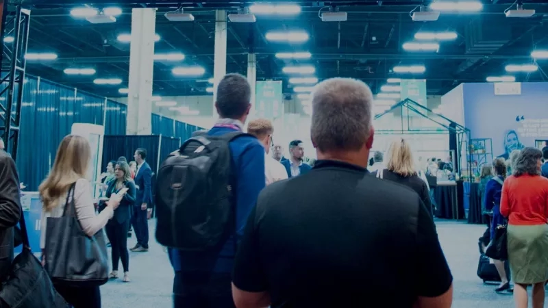 A group of people are at a tradeshow with many booths and a bright blue wall behind them.