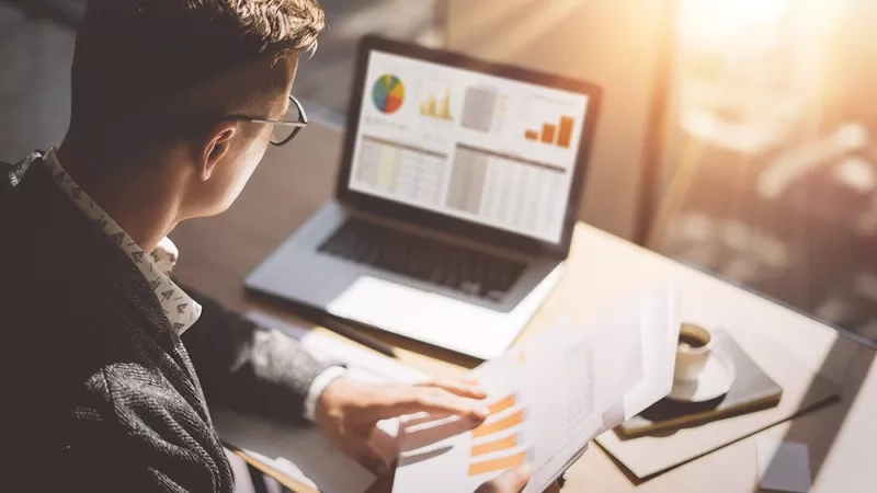 Businessman with budget papers in front of laptop with charts and graphs on screen.