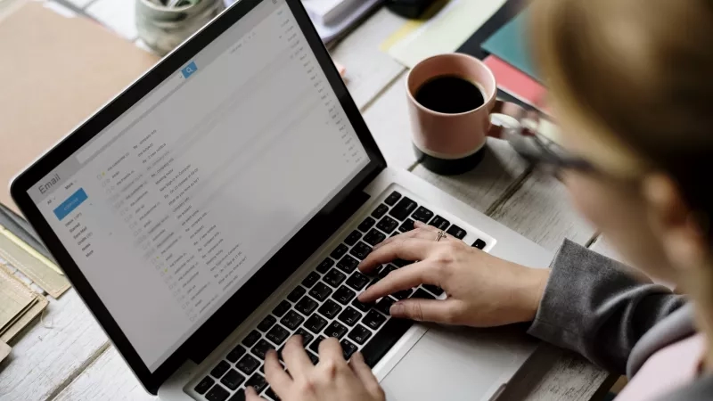 Woman typing on a laptop with an email open in front of her on a desk.