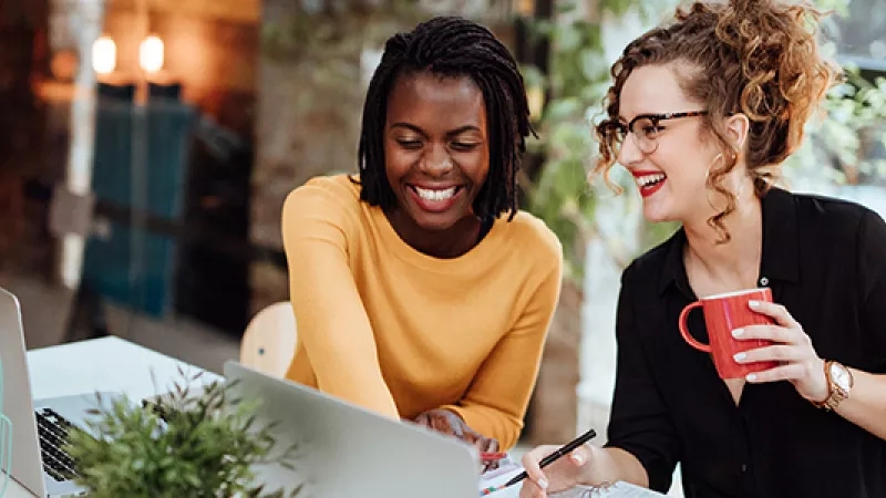 Two women having a meeting, one holding a coffee mug, and the other working on a laptop.