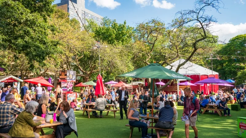 Crowded outdoor cafe with tents and tables, surrounded by trees and buildings under a clear sky.