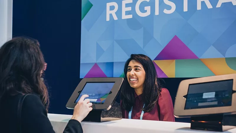 Woman registering for an event on a digital kiosk, while another woman is standing and smiling.