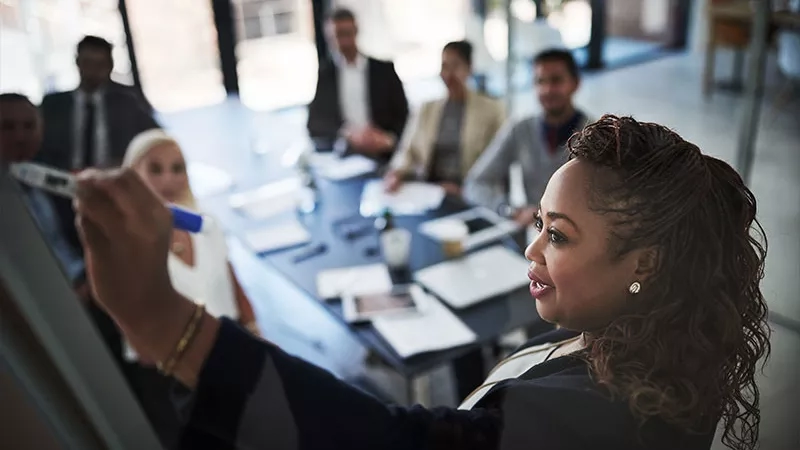 Woman writing on a white board in front of a group of people in a meeting room.