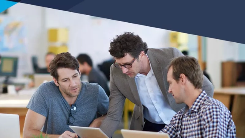 Three men in an office discuss the five myths of social media marketing on laptops and tablets.