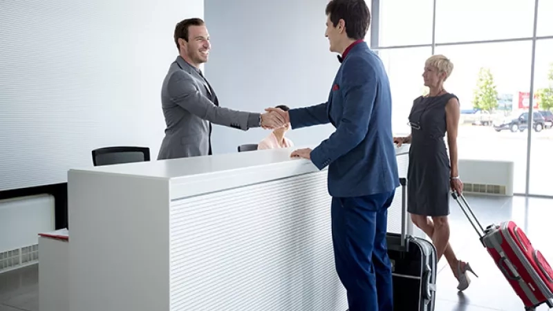 A man greets a guest at the front desk of a hotel while another guest walks by with luggage.
