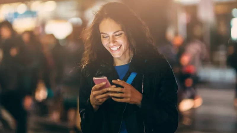 A woman is smiling while using her phone, surrounded by blurred people in an urban night scene.