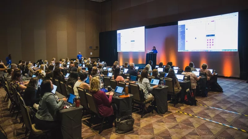 A conference hall where a woman is giving a presentation and the audience is watching on their laptops.