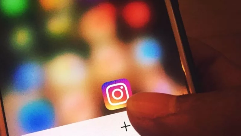 A hand holding a phone with Instagram on the screen and a blurred background of colorful lights.