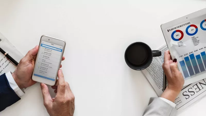 Two hands holding a tablet and smartphone in front of a newspaper and coffee cups.
