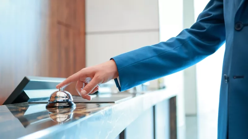 Person in blue jacket ringing a silver bell at a hotel front desk.