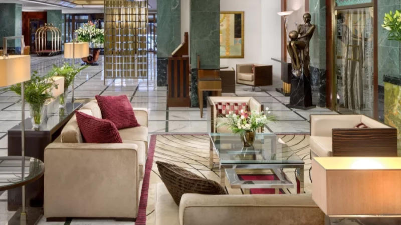 The lobby of the Hotel Alcron features couches, a glass table, and a carpet on the floor.