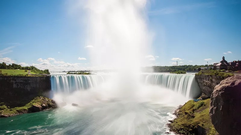 The Niagara Falls cascades over the cliff, creating a breathtaking view with the blue sky and greenery.