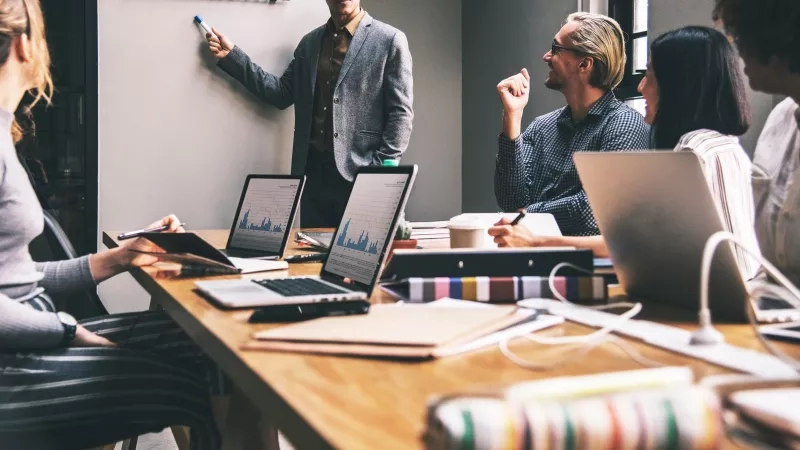 A team of adults brainstorming and analyzing in a conference room with laptops, notebooks, and a whiteboard.