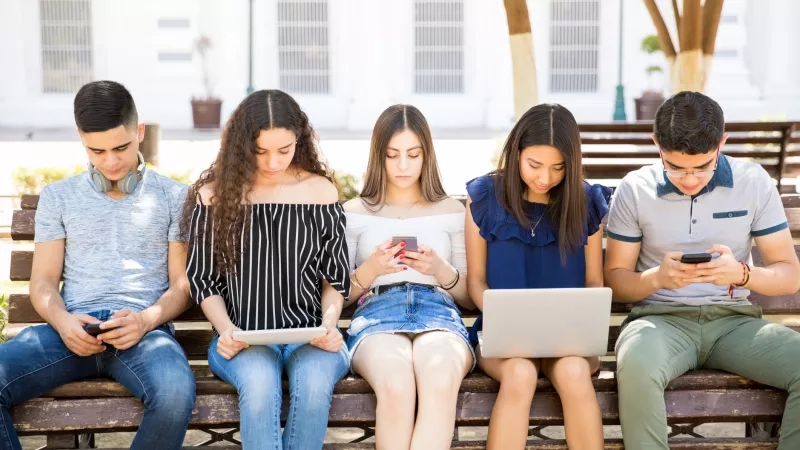Group of young people using technology devices in park, representing the digital lifestyle of Gen Z.