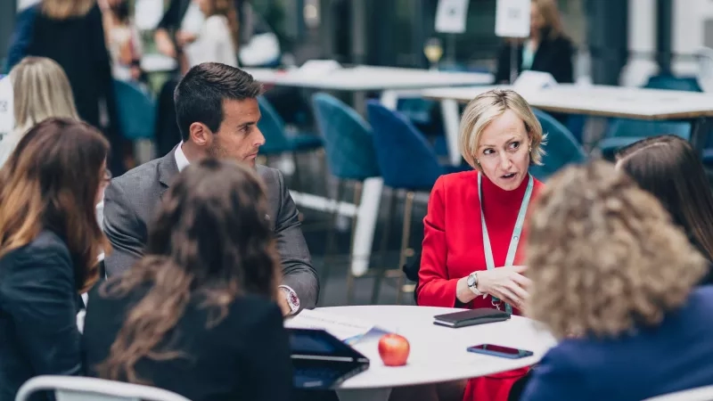 Group of people at a table with a lady in red talking and others listening at an event.